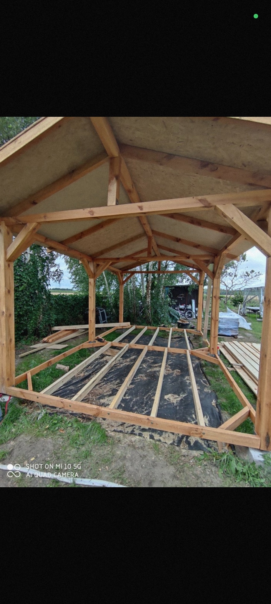 Konstrukcja drewnianej altany w trakcie budowy; widoczne elementy szkieletu dachu i podłogi na czarnej macie. Tło: zieleń ogrodu, częściowo zadaszone miejsce.