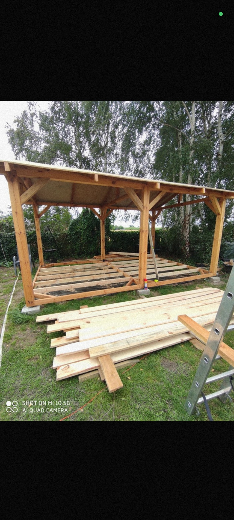 Drewniana konstrukcja z zadaszeniem w trakcie budowy na zewnątrz. Widoczne deski, drabina i narzędzia. Tło stanowią drzewa i zieleń. Ujęcie z dołu.
