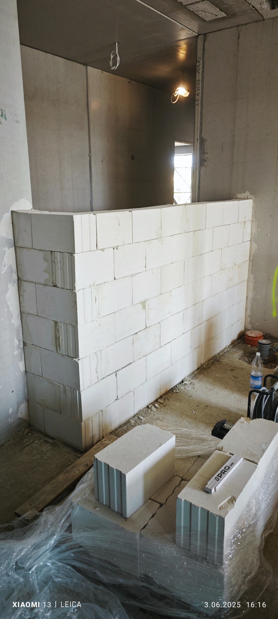 Wnętrze w trakcie budowy z częściowo wymurowaną ścianką działową z białych bloczków, poziomica leży na bloczkach. Widoczne surowe ściany i podłoga.