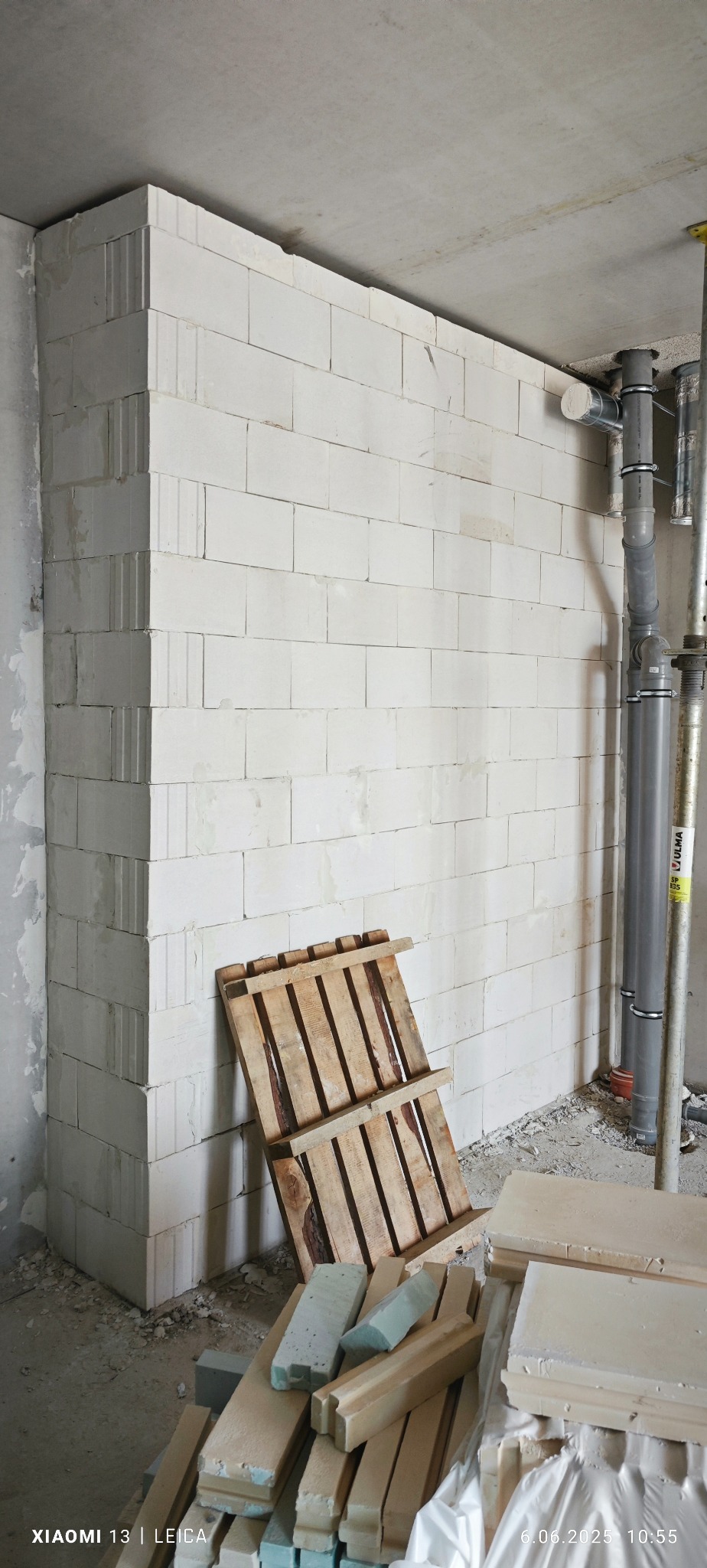 Wzniesiona ściana z białych bloczków betonu komórkowego w surowym wnętrzu, wsparta drewnianą paletą. Widoczne rury instalacyjne i resztki materiałów budowlanych.