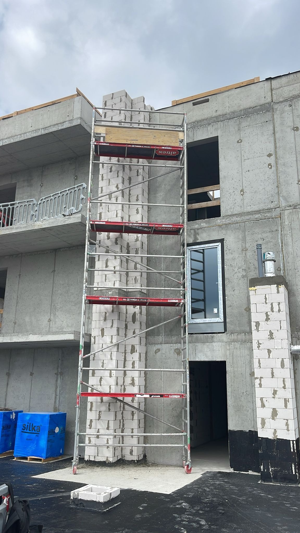 Wznoszenie ściany z białych bloczków silikatowych, widoczne rusztowanie i okno. Budowa w toku, surowy stan budynku. Na pierwszym planie opakowania bloczków.