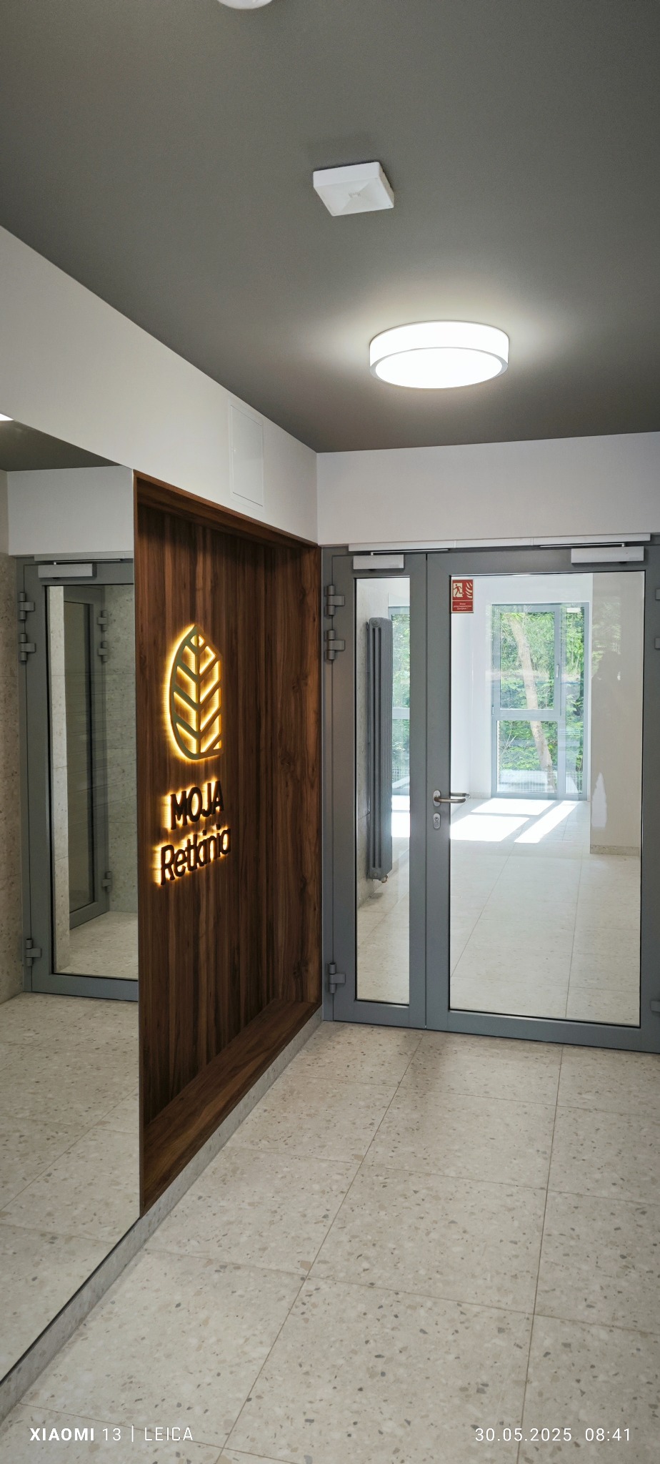 Nowoczesne wykończenie wnętrza: drewniana ścianka z podświetlanym logo 'MOJA Retkinia' w korytarzu z przeszklonymi drzwiami i jasną podłogą z płytek.