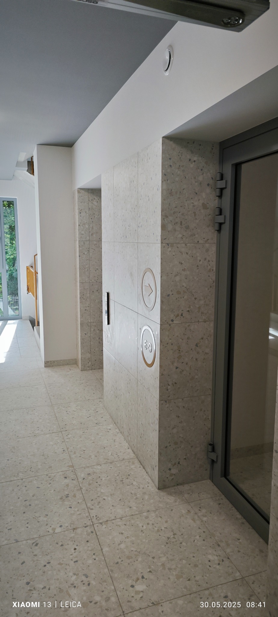 Beżowa posadzka i ściana z płytek lastryko w korytarzu budynku. Widoczny panel sterowania windy z przyciskami i strzałką. Szare drzwi wejściowe z zawiasami.