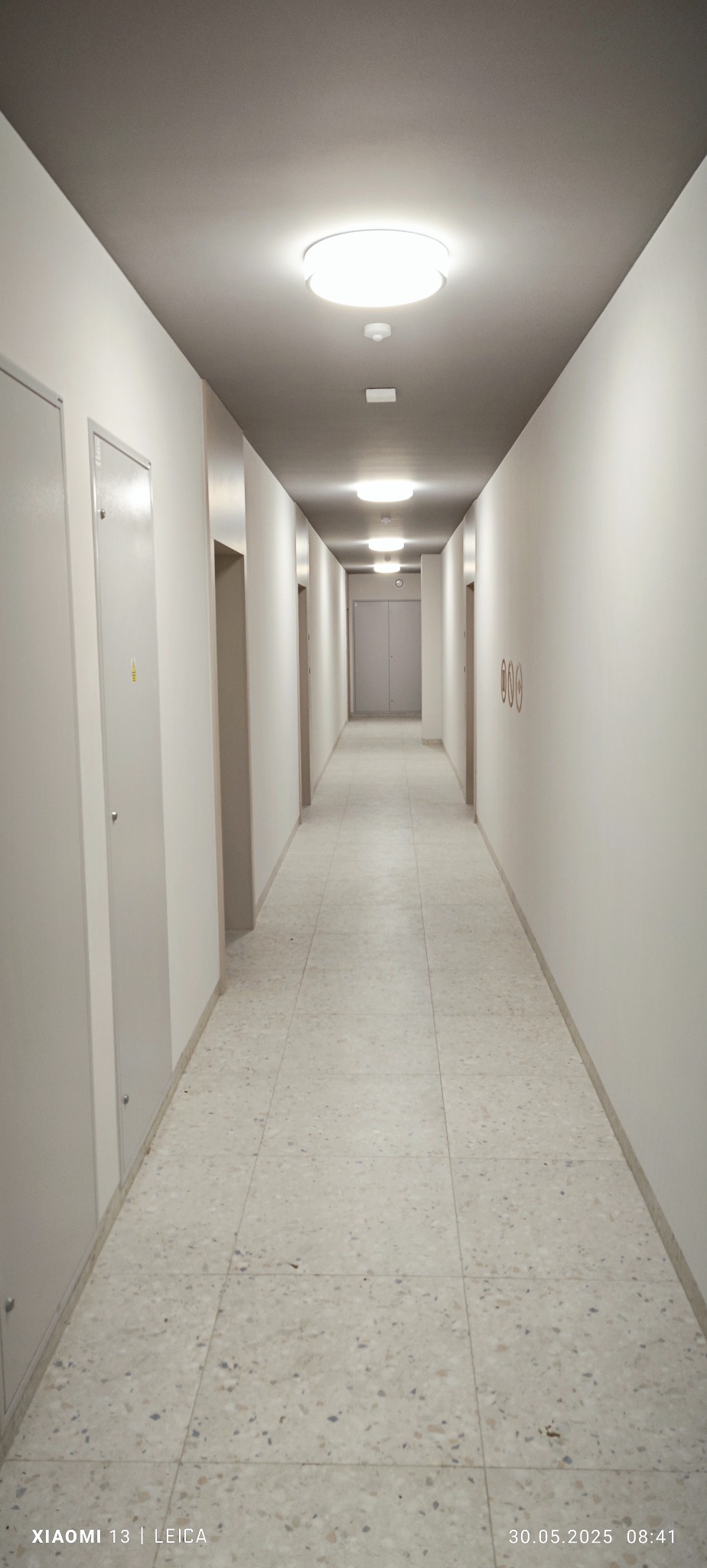 Długi, jasny korytarz z terakotą w budynku mieszkalnym. Widoczne drzwi po bokach i oświetlenie sufitowe. Minimalistyczny design i neutralna kolorystyka.