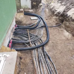 Instalacja rur w wykopie przy budynku, czarne rury karbowane i gładkie, czerwone złączki, ziemia, widok z góry.