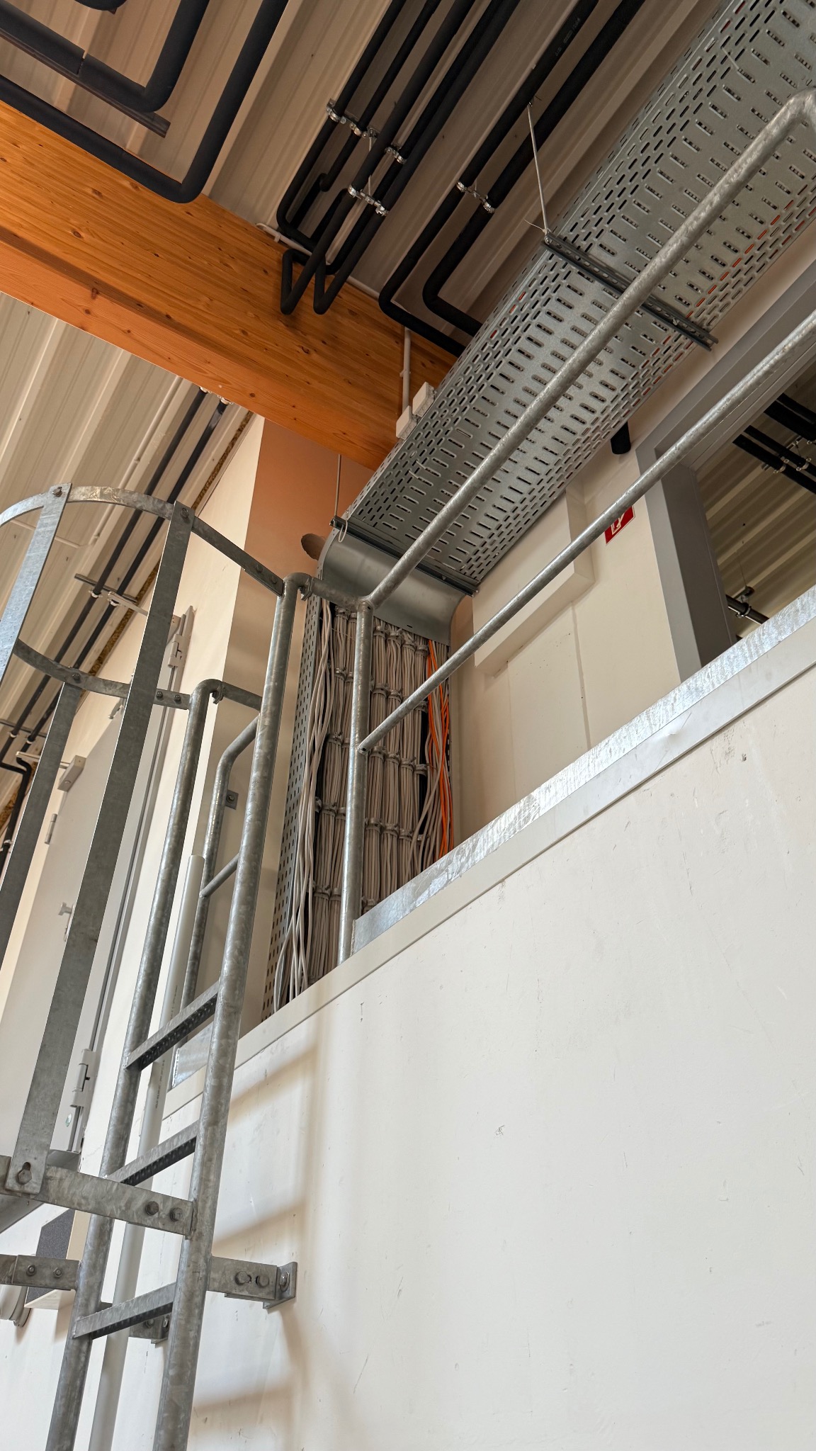 Metalowa drabina, koryto kablowe z przewodami i rury instalacyjne pod sufitem w hali. Widok z dołu, kable uporządkowane w korycie.