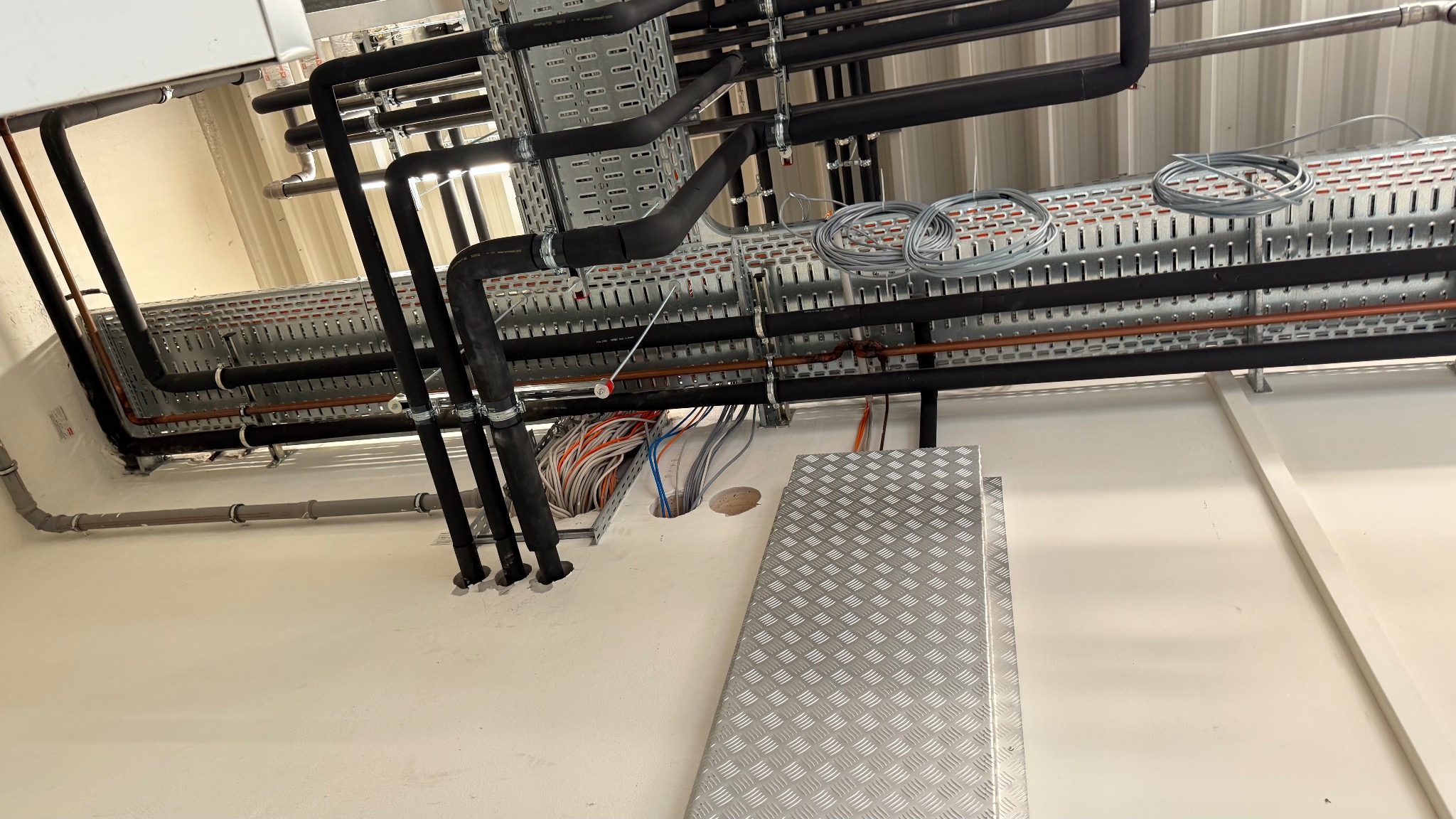 Widok na sufit z korytkami kablowymi, rurami i przewodami. Metalowa platforma pod sufitem. Kolorowe kable w otworze w suficie.