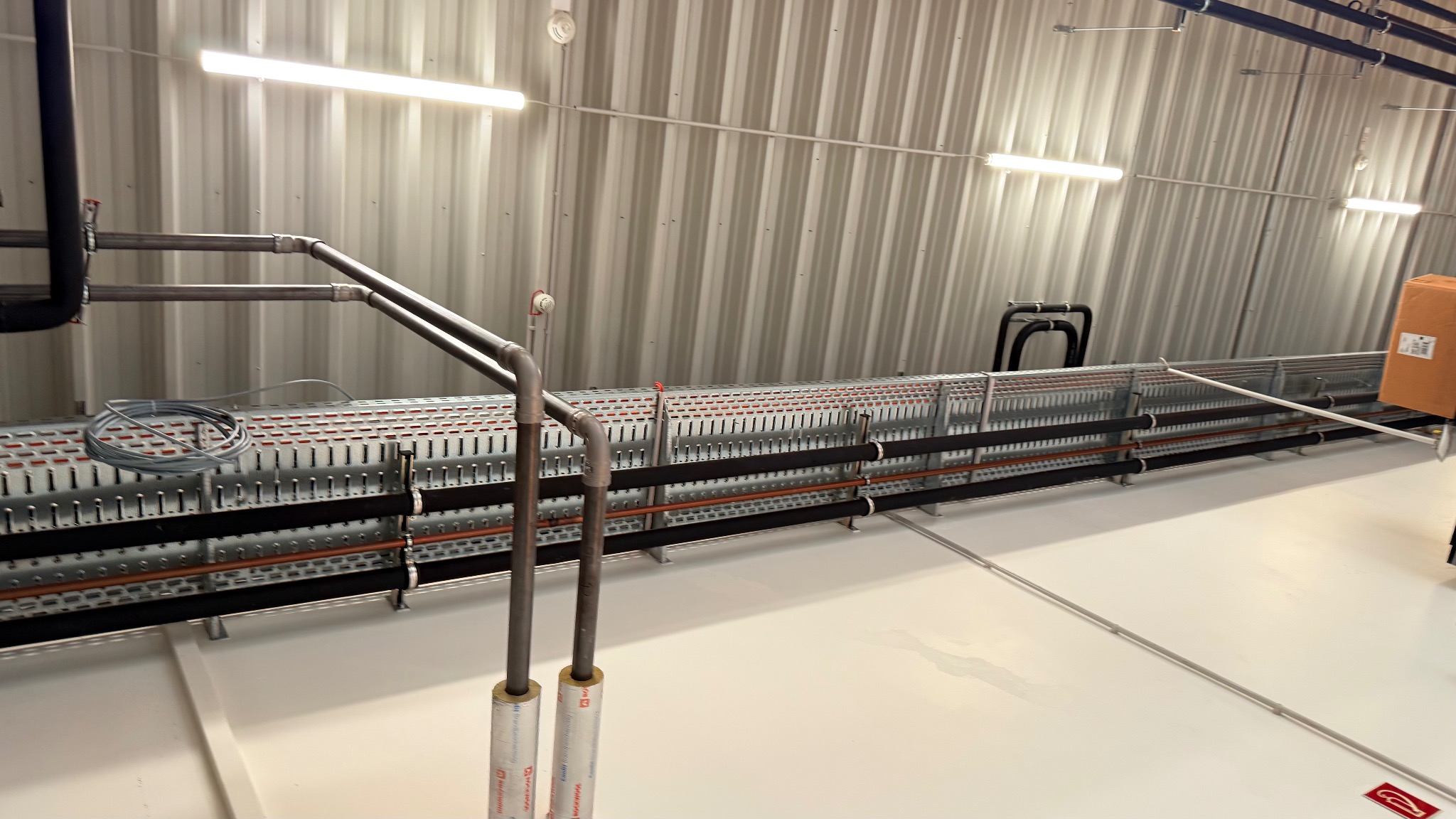 Długa trasa kablowa z rurami i przewodami, zamontowana pod sufitem w hali z blachy falistej. Widoczne elementy instalacji i oświetlenie.