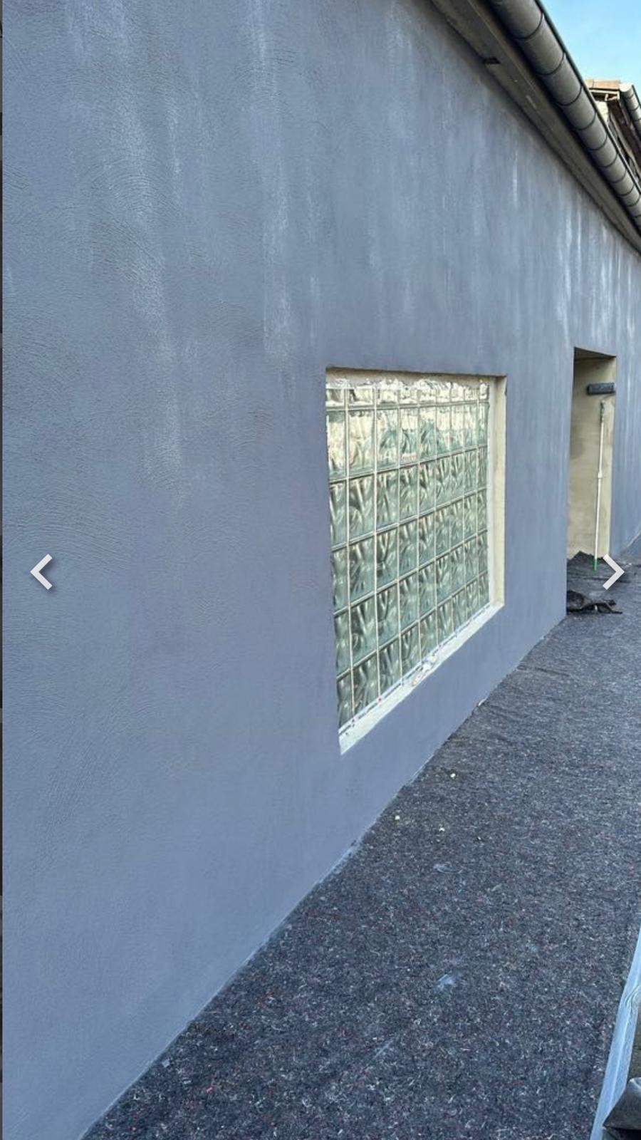 Świeżo otynkowana szara ściana zewnętrzna z oknem z luksferów. Widoczny wałek malarski w otworze drzwiowym i zabezpieczona podłoga.