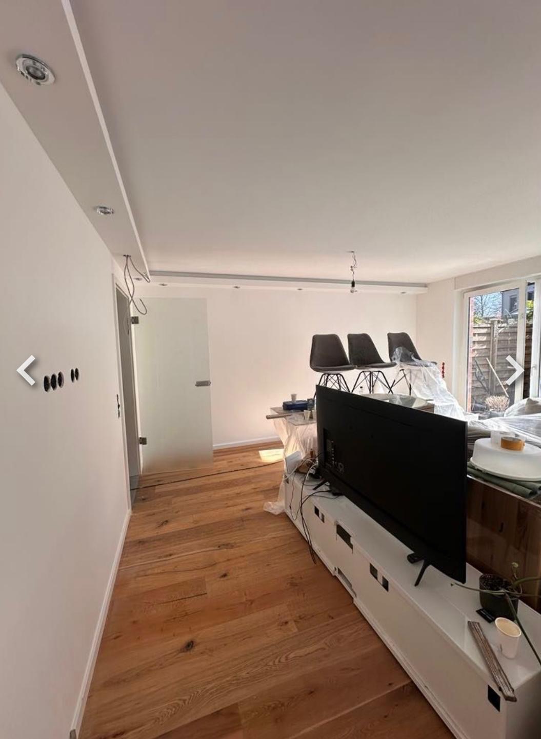 Jasne wnętrze po remoncie: świeżo pomalowane ściany, drewniana podłoga, biała listwa sufitowa z oświetleniem punktowym. W tle meble zabezpieczone folią.