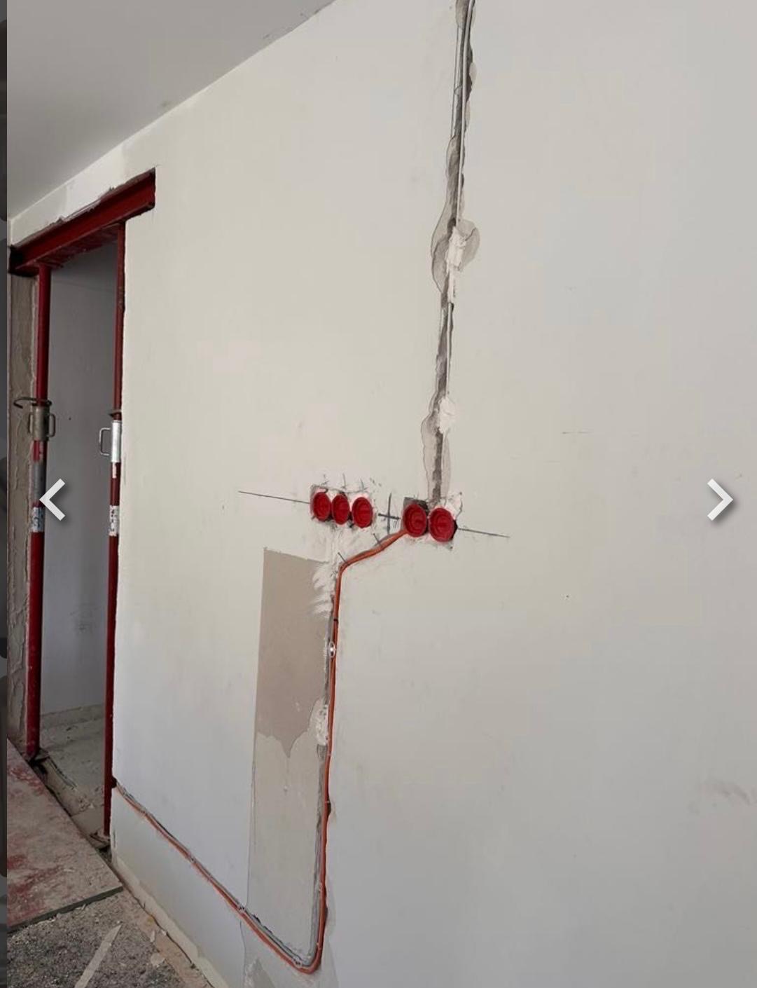 Ściana z bruzdami pod kable i puszkami elektrycznymi w kolorze czerwonym. Widoczny przewód elektryczny poprowadzony wzdłuż ściany. W tle ościeżnica drzwiowa.