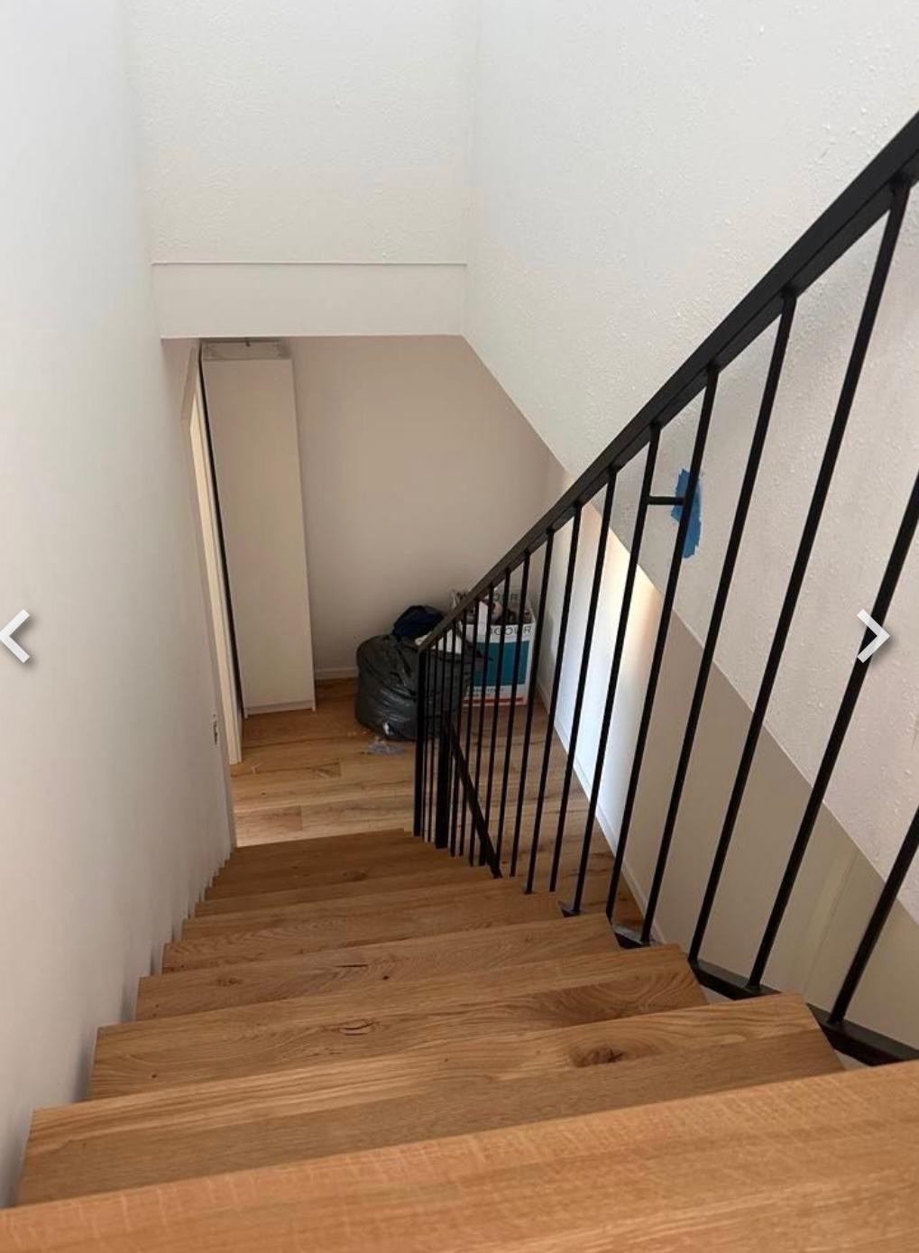 Drewniane schody z czarną balustradą w jasnym wnętrzu. Widok z góry na dół, ukazujący detale wykończenia i materiałów. W tle widoczny worek na śmieci.