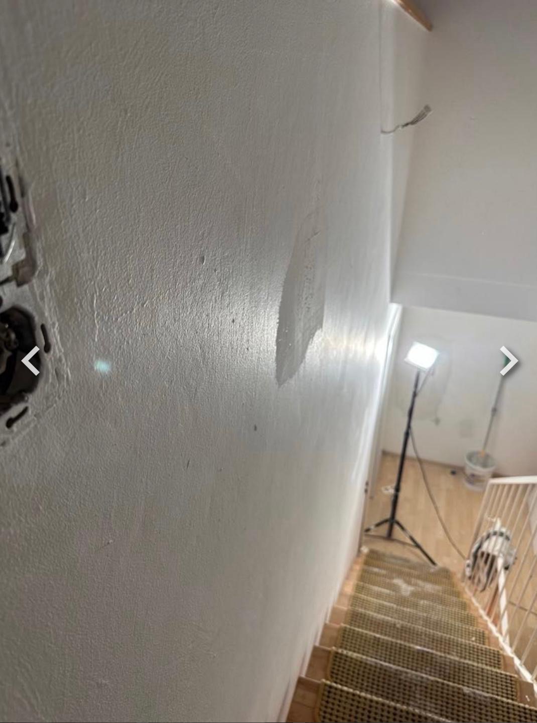 Ściana po szpachlowaniu z widocznym gniazdkiem elektrycznym w trakcie remontu klatki schodowej. Na podłodze widoczny reflektor budowlany i schody.