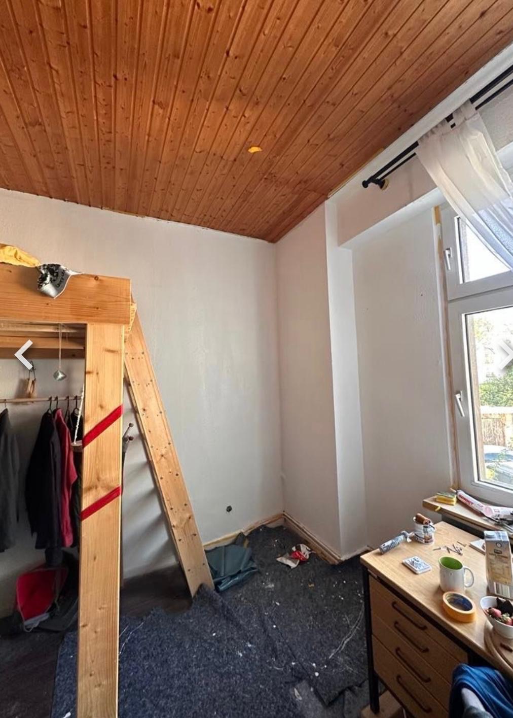 Pokój w trakcie malowania: drewniany sufit, świeżo pomalowane ściany na biało, okno z firanką, meble i drabina. Widoczne ślady farby na podłodze.