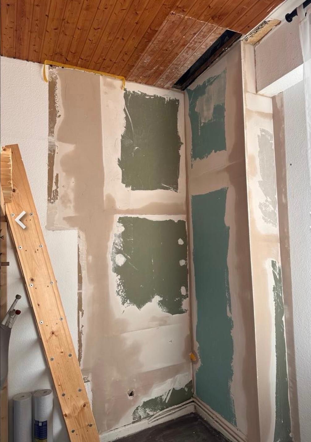 Ściana w trakcie szpachlowania z widocznymi łatami i drewnianym sufitem. Fragment drabiny oparty o ścianę. Prace wykończeniowe w pomieszczeniu.