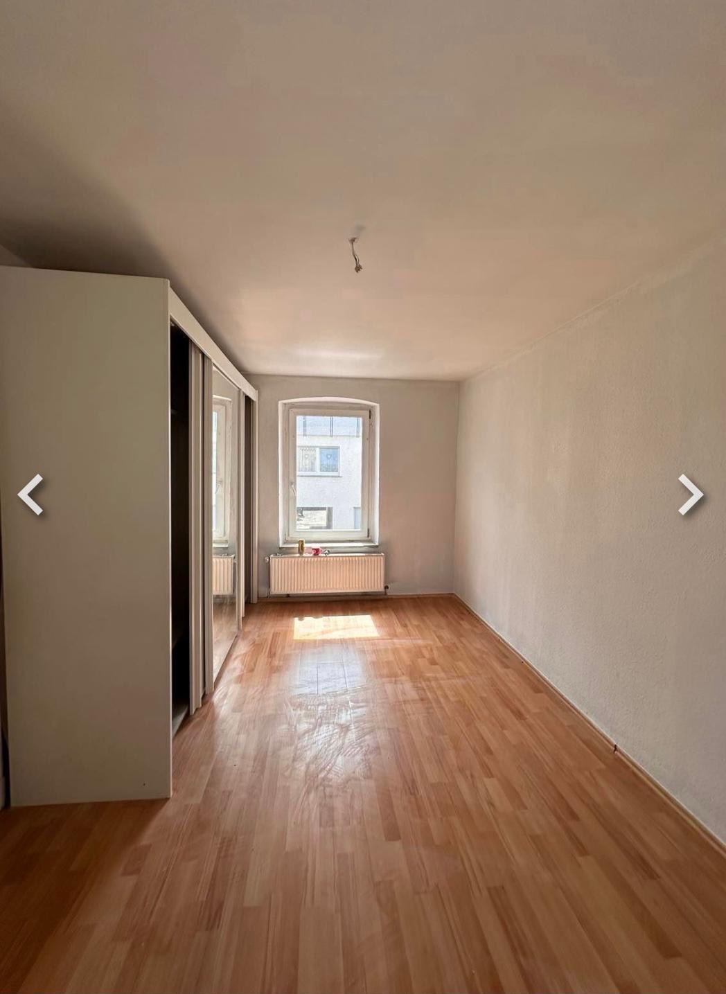 Pusty pokój po remoncie: parkiet, białe ściany, okno z grzejnikiem i szafa z lustrami. Widoczne ślady po lampie na suficie. Jasne, słoneczne wnętrze.