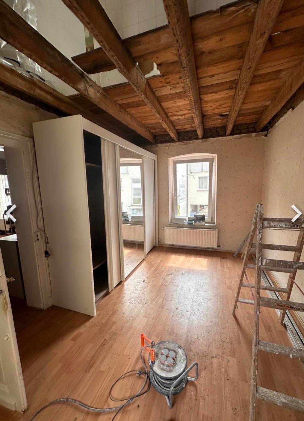 Pokój w trakcie remontu: odsłonięty drewniany strop, stara podłoga, drabina i sprzęt do szlifowania podłogi. Widoczna szafa z lustrami i okno z grzejnikiem.