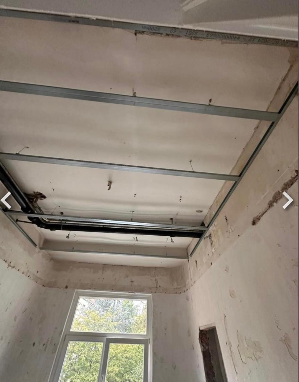 Stelaż sufitu podwieszanego z profili metalowych w trakcie montażu. Widoczne przewody instalacji nad stelażem. W tle okno z widokiem na zieleń.