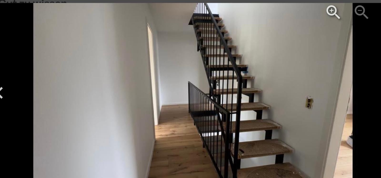 Wnętrze domu z drewnianą podłogą i schodami z czarną metalową balustradą. Schody zabezpieczone kartonem. Widoczny fragment ściany z gniazdkiem elektrycznym.