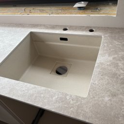 BOGUSŁAW STRYJAK ARTWOOD PRACOWNIA - Beżowy zlew wkomponowany w blat imitujący marmur, widoczne otwory na baterię i dozownik, fragment szafki kuchennej. Ujęcie z góry.