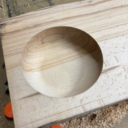 BOGUSŁAW STRYJAK ARTWOOD PRACOWNIA - Płyta z jasnego drewna z wyfrezowanym okrągłym wgłębieniem, otoczona wiórami. Widoczne pomarańczowe podkładki. Obróbka CNC w toku.