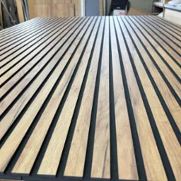 Panel akustyczny z drewnianymi lamelami w Odolanowie. Dekoracyjny element wykończeniowy z wyraźnym rysunkiem drewna i czarnym filcem w tle, ułożony na płasko.