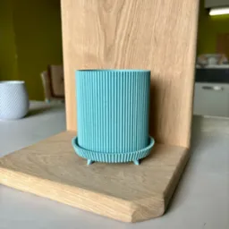 Turkusowy, prążkowany wazon z podstawką, wydrukowany w technologii 3D, umieszczony na drewnianej półce na tle zielonej ściany i wyposażenia kuchni.