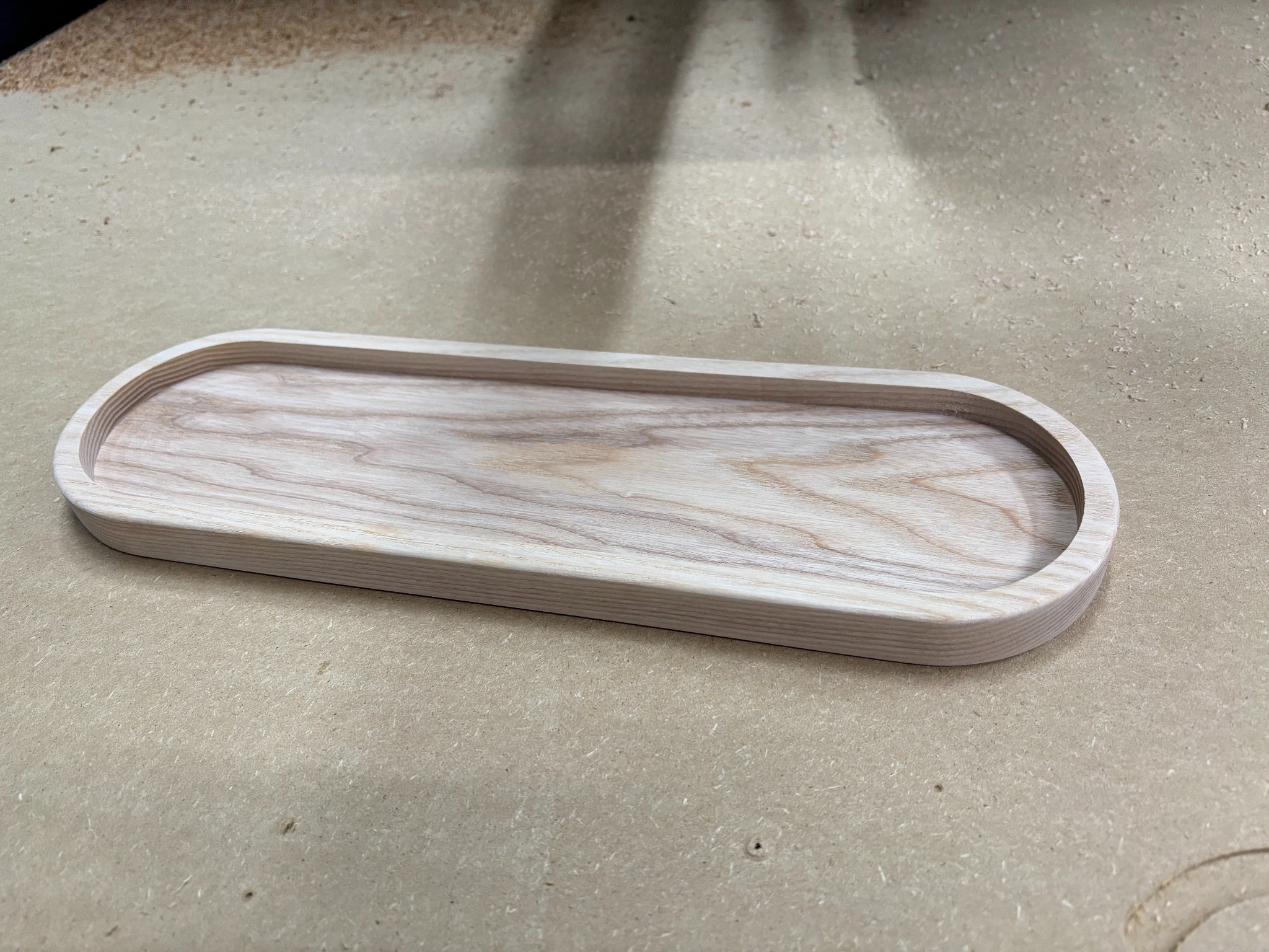 Owalna, jasna taca z drewna na stole warsztatowym, wykonana z frezowanego drewna. Widoczne ślady obróbki i wióry. Minimalistyczny design.