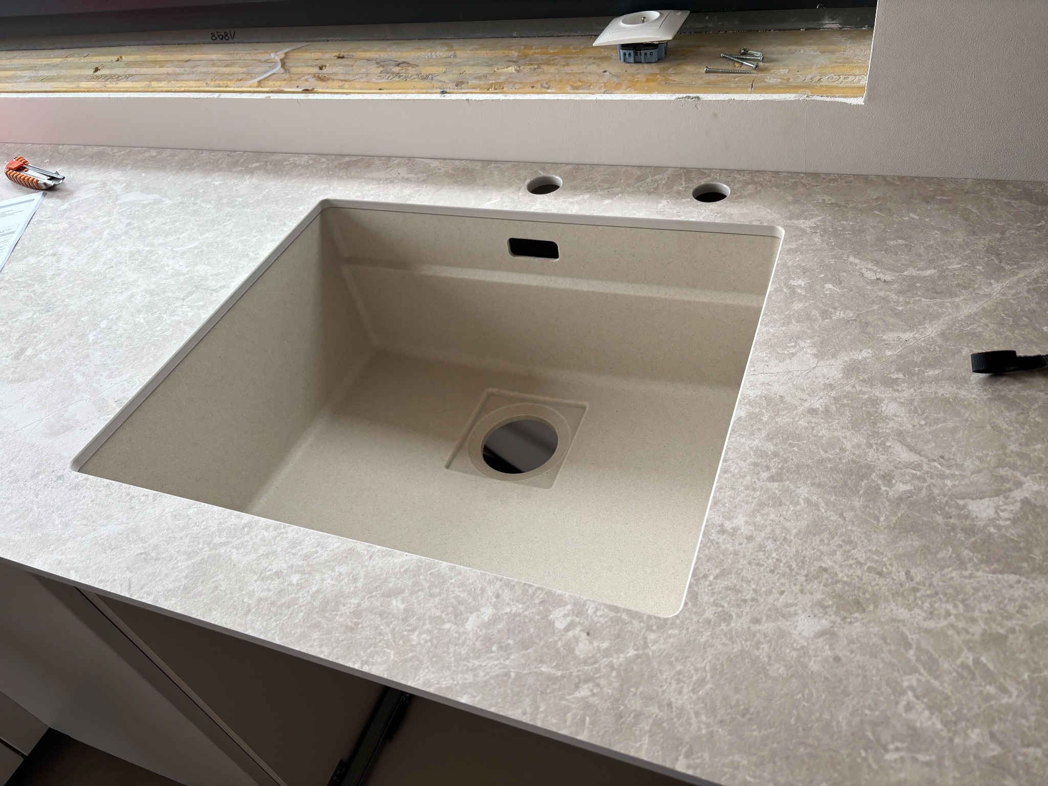 Beżowy zlew wkomponowany w blat imitujący marmur, widoczne otwory na baterię i dozownik, fragment szafki kuchennej. Ujęcie z góry.