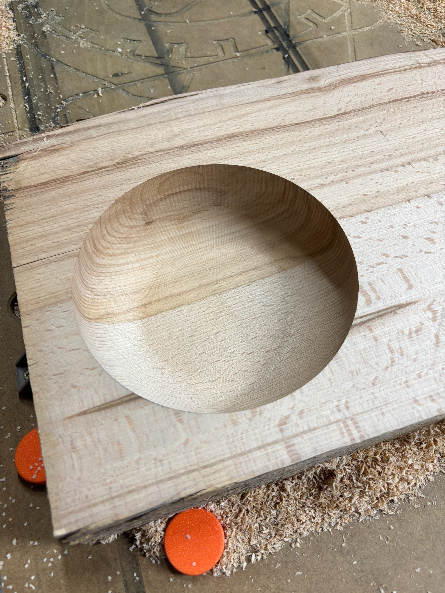 Płyta z jasnego drewna z wyfrezowanym okrągłym wgłębieniem, otoczona wiórami. Widoczne pomarańczowe podkładki. Obróbka CNC w toku.