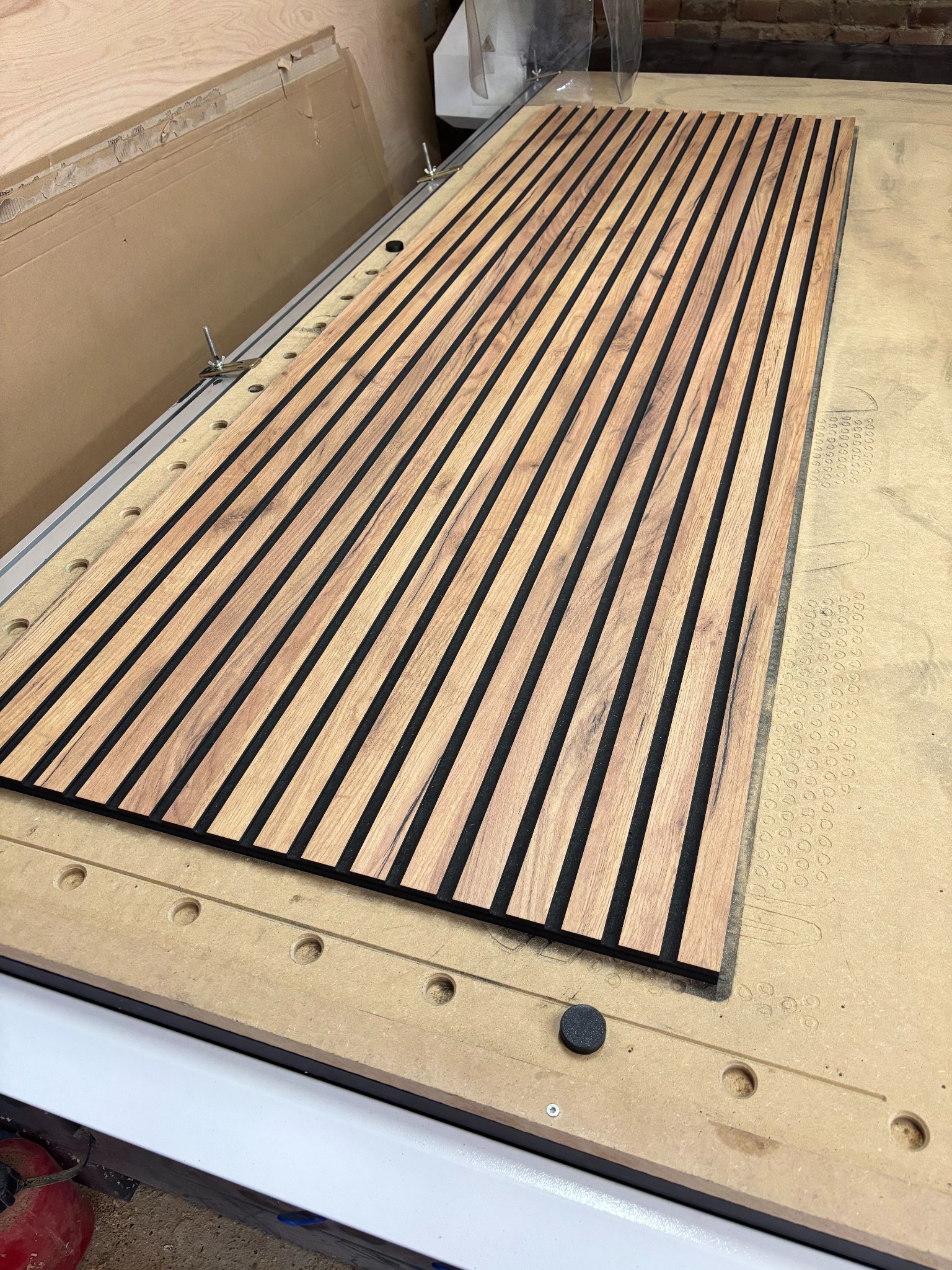 Panel lamelowy z drewna i czarnego filcu na stole frezarki CNC, widoczne mocowania. Detal obróbki, bliski kadr, ukośne ułożenie lameli.