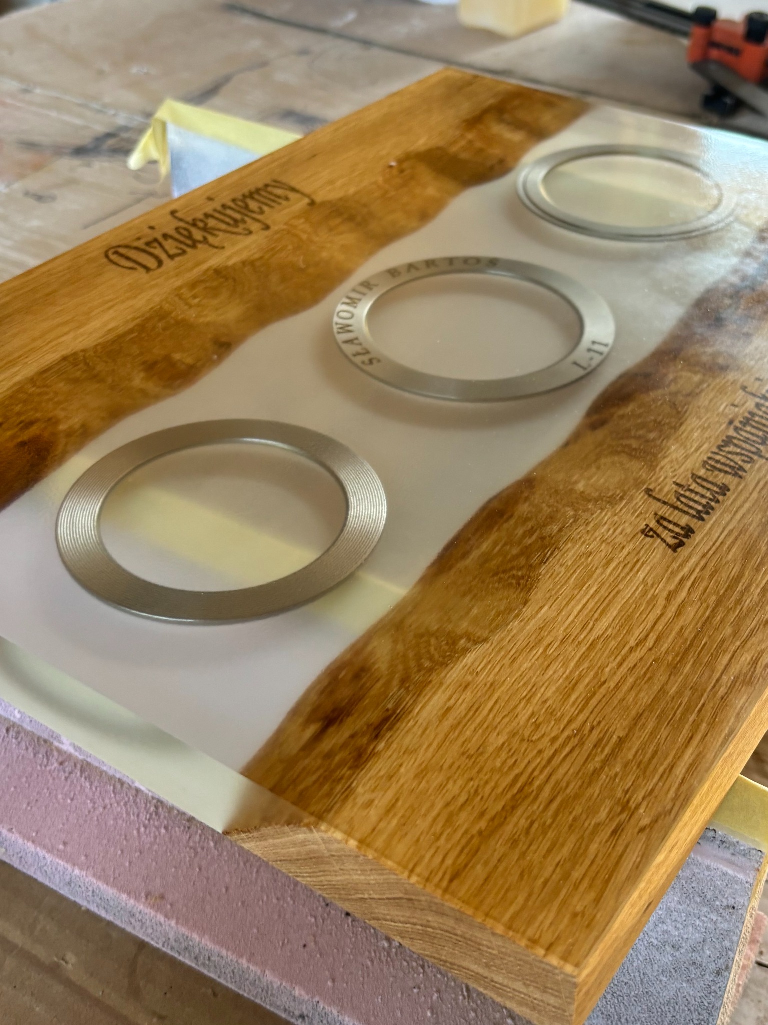 Deska z drewna dębowego z żywicą epoksydową, wtopione metalowe obręcze i wygrawerowane napisy. Blat na etapie wykończenia, zabezpieczony taśmą malarską.