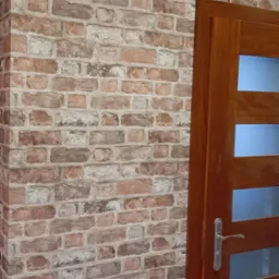 Ściana z cegły dekoracyjnej w odcieniach brązu i bieli, przylegająca do drewnianych drzwi z przeszkleniami, tworzy rustykalny, ale nowoczesny wygląd wnętrza.