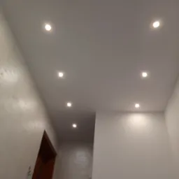 Nowoczesny sufit podwieszany z oświetleniem punktowym w korytarzu, ściany wykończone w jasnych odcieniach, widoczny fragment ościeżnicy drzwiowej.