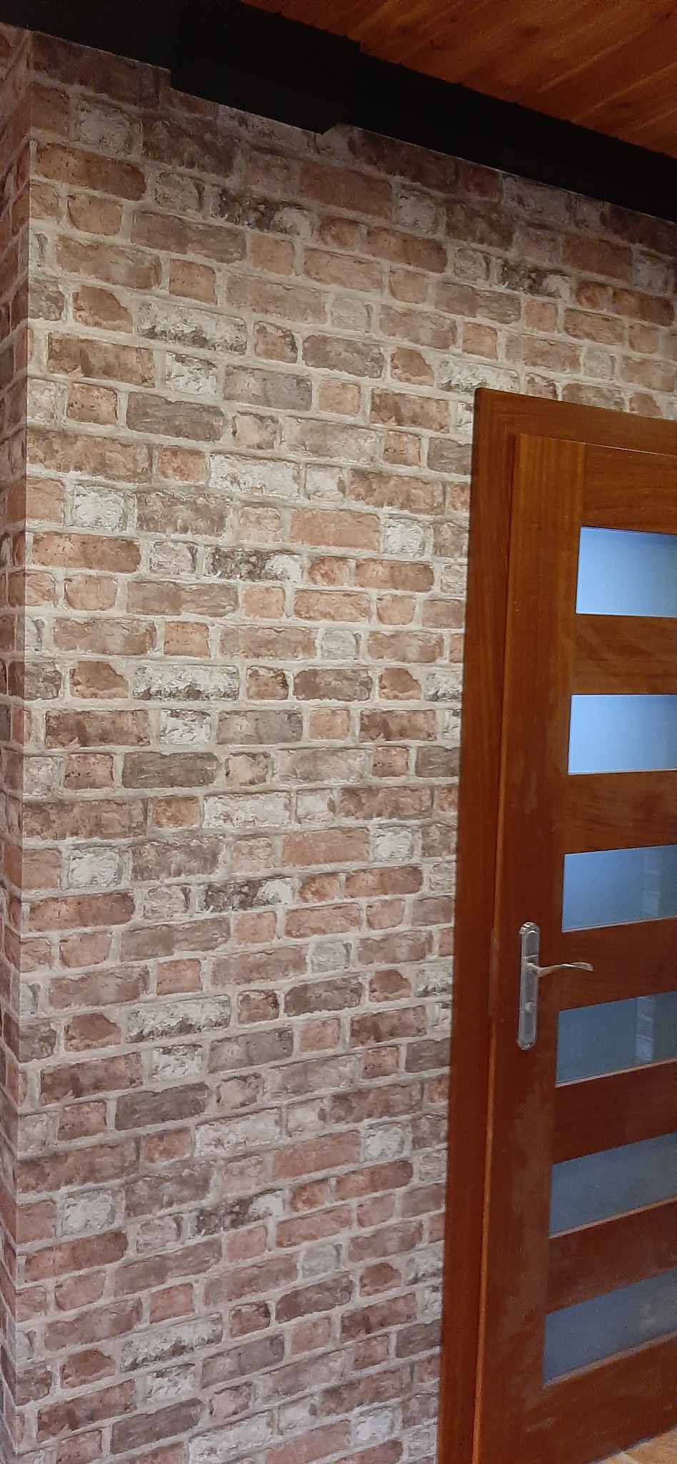 Ściana z cegły dekoracyjnej w odcieniach brązu i bieli, przylegająca do drewnianych drzwi z przeszkleniami, tworzy rustykalny, ale nowoczesny wygląd wnętrza.