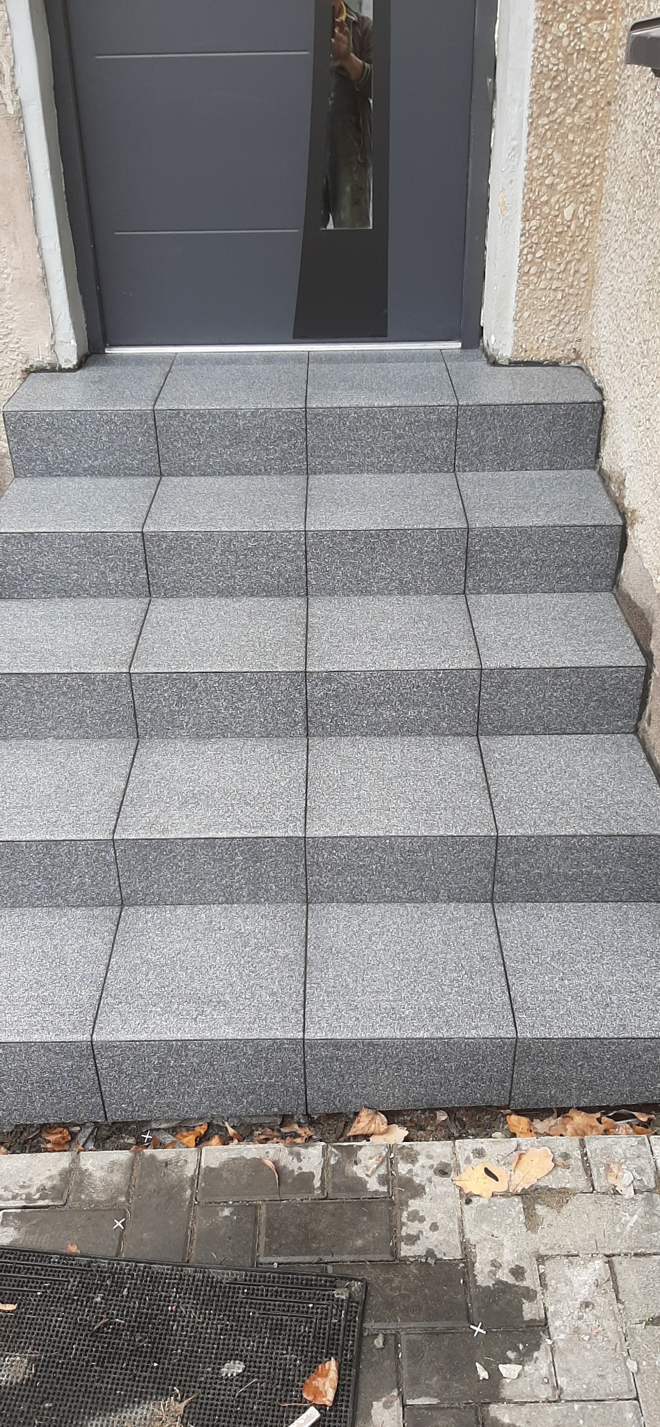 Wykończone schody wejściowe z szarych, kwadratowych płytek, prowadzące do szarych drzwi z szybą. Widoczne detale fug i faktura płytek.