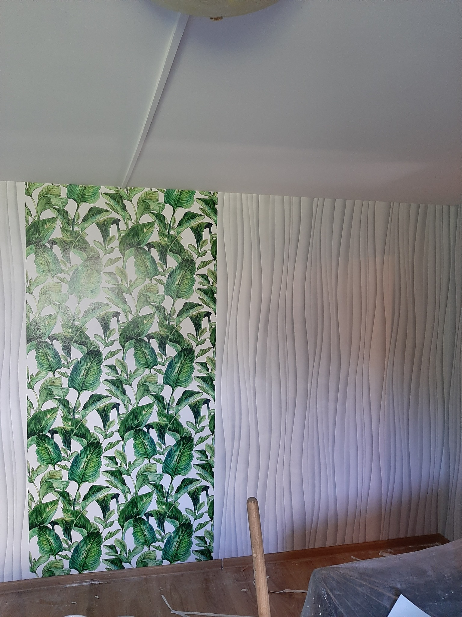 Fragment ściany: połowa z tapetą w zielone liście, druga z białą, falistą strukturą. Widoczny fragment sufitu i podłogi. W tle narzędzia malarskie.
