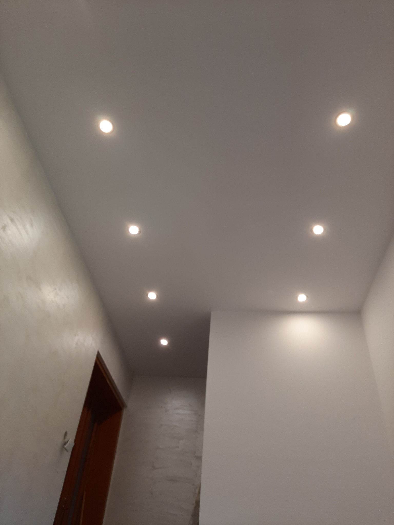 Nowoczesny sufit podwieszany z oświetleniem punktowym w korytarzu, ściany wykończone w jasnych odcieniach, widoczny fragment ościeżnicy drzwiowej.