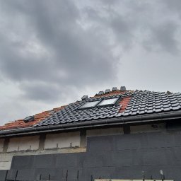 Zarko sp. z o.o. - Montaż dachówek ceramicznych na dachu z oknami połaciowymi, widoczne rusztowania i ocieplenie budynku styropianem grafitowym. Pochmurne niebo.