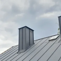 Szary dach z blachy na rąbek stojący z kominem i oknem dachowym, widok pod kątem. Pochmurne niebo w tle, podkreślające nowoczesny styl.