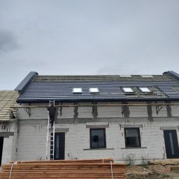 Zarko sp. z o.o. - Montaż ciemnej dachówki na nowym domu z oknami dachowymi. Na drabinie dekarz, w tle szare niebo. Na dole drewniane belki.