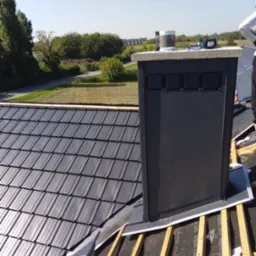 Ciemnoszara dachówka ceramiczna na połaci dachu w trakcie montażu, widoczny komin wentylacyjny i elementy konstrukcji drewnianej. Słoneczny dzień.