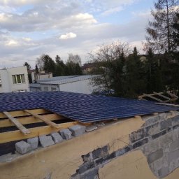 Zarko sp. z o.o. - Montaż ciemnej blachy trapezowej na dachu budynku. Widoczne drewniane elementy konstrukcyjne i fragmenty muru. Tło stanowią budynki i drzewa.