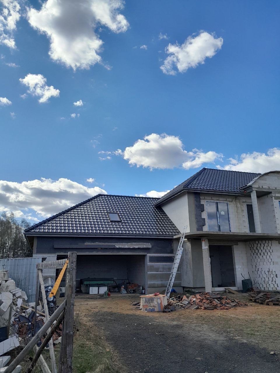 Dom w trakcie budowy, z widocznym dachem pokrytym ciemną dachówką i elementami konstrukcyjnymi. Na pierwszym planie gruz i drabina.
