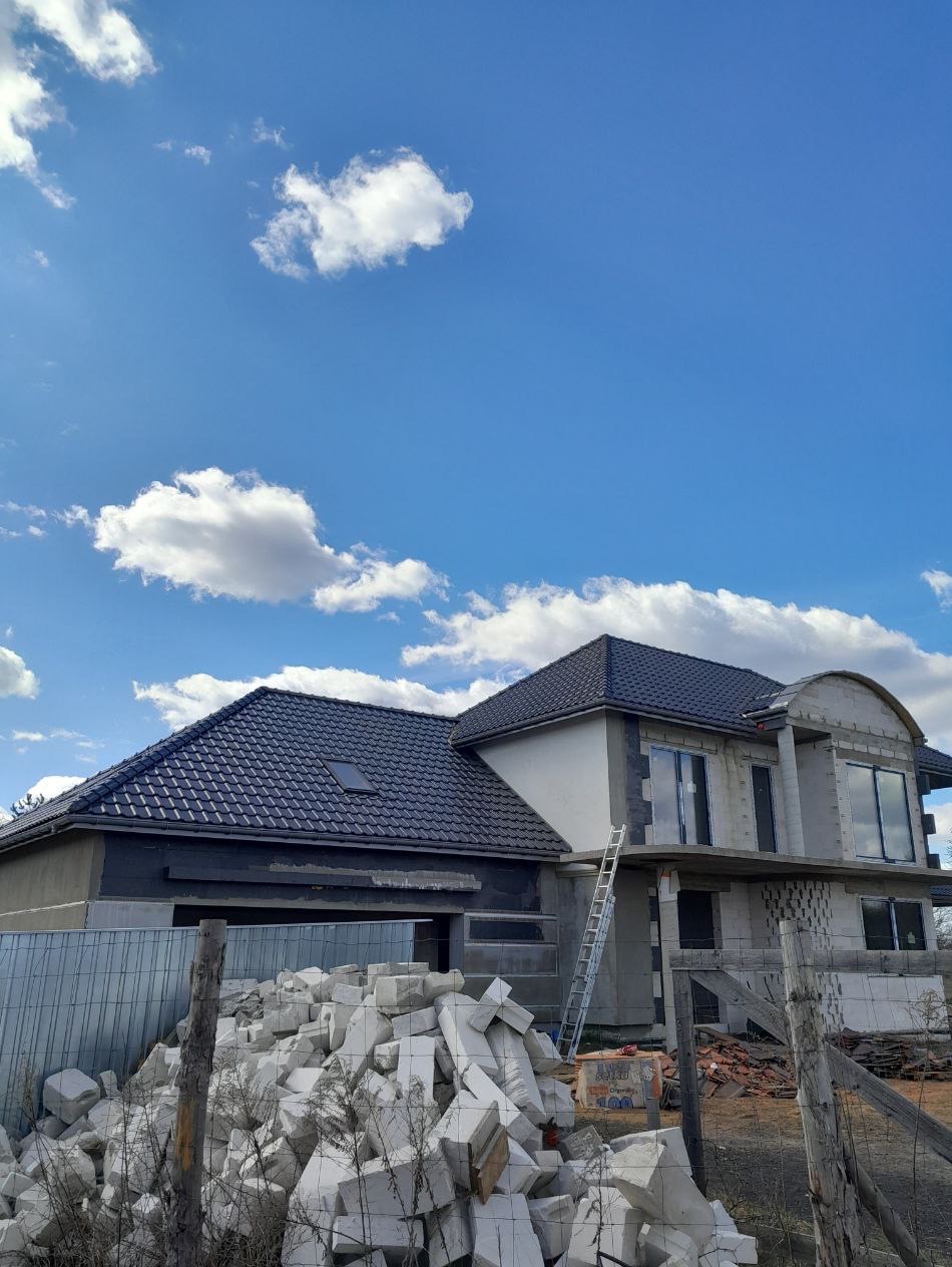 Dom w trakcie budowy z ciemną dachówką i stertą białych pustaków. Widoczne okna, drabina oparta o budynek na tle błękitnego nieba z chmurami.