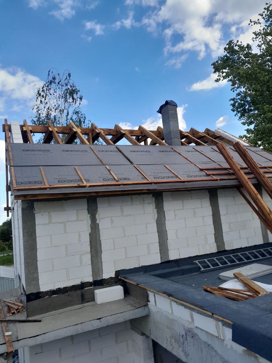 Budowa dachu z drewnianą konstrukcją i membraną. Widoczny komin i białe bloczki konstrukcyjne. Niebieskie niebo w tle. Detal budowy dachu.