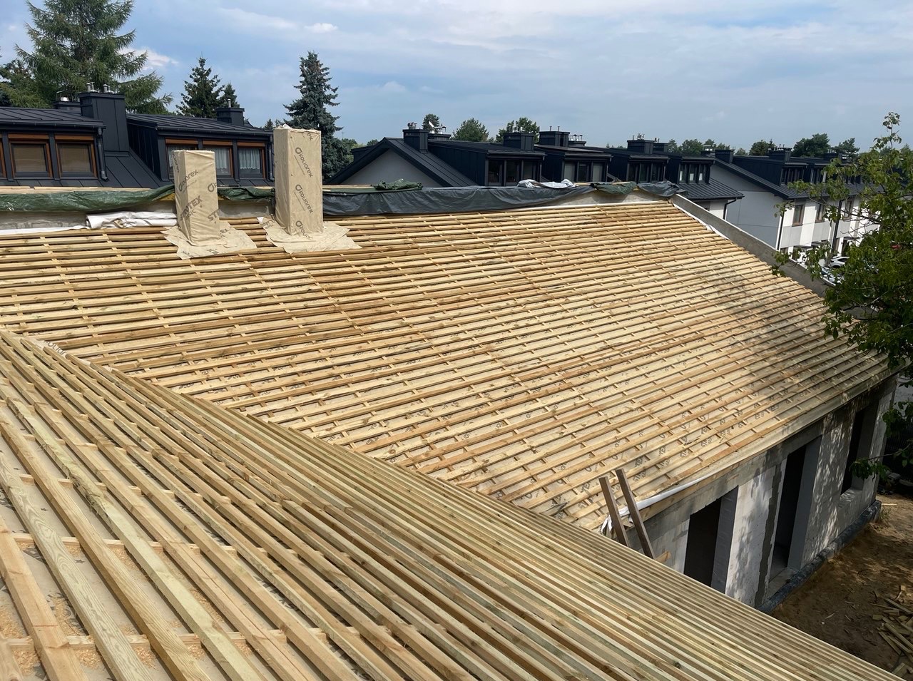 Dach w trakcie budowy: drewniana konstrukcja łat na budynku z czarnymi kominami w tle. Widoczne zabezpieczone kominy i fragment drabiny.