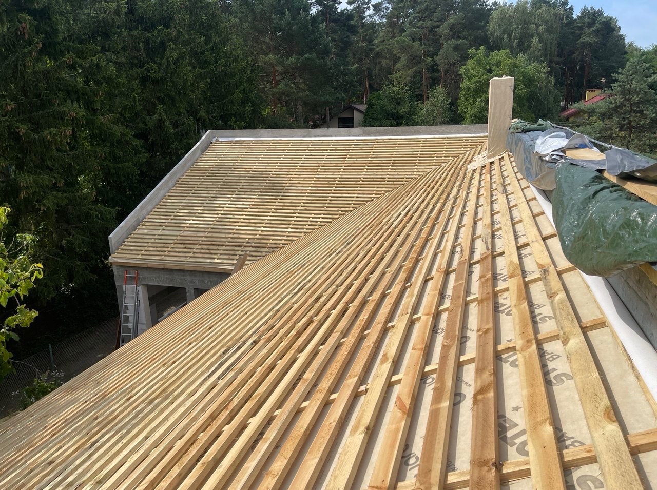 Wysoki kąt na konstrukcję dachu z drewnianymi łatami, widoczne elementy komina i częściowo pokrycie papą. W tle drzewa i budynek.