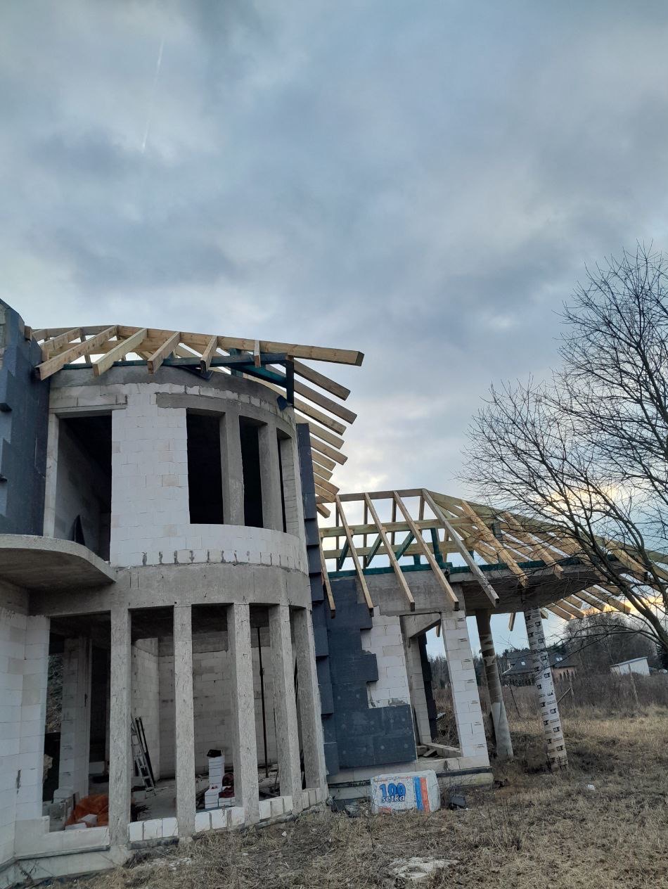 Surowy stan budynku z drewnianą konstrukcją dachu w trakcie budowy, widoczne bloczki i ocieplenie, pochmurne niebo w tle, drzewo bez liści po prawej stronie.