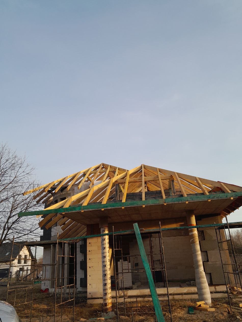 Budowa domu jednorodzinnego w trakcie realizacji: widoczna konstrukcja dachu z drewnianych krokwi, ściany z pustaków i rusztowania. Niebo w tle.